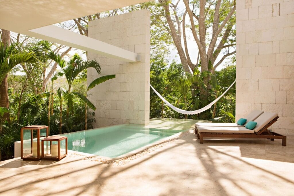 Outdoor pool area with a hammock, lounge chair, and nested tables, surrounded by lush tropical greenery and stone walls reminiscent of Yucatan haciendas.