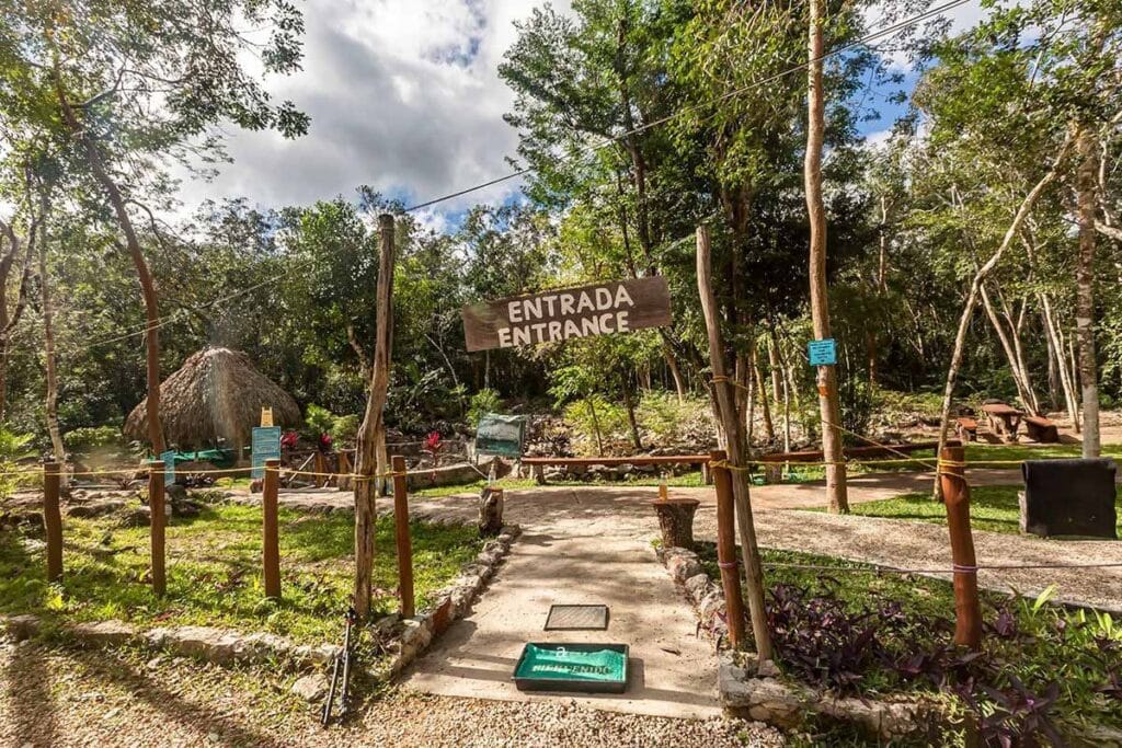 A rustic entrance sign reads "Entrada" amidst lush greenery, with a traditional hut visible in the background, indicating a natural or cultural park.