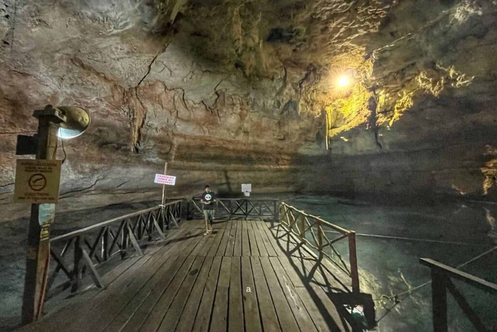 A person stands on a wooden walkway inside a large, illuminated cave with high ceilings and rocky walls. Signs are visible nearby.