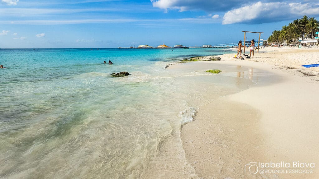 Sunny beach scene with turquoise water, several people enjoying the shore, and palm trees lining the sand. Clear blue skies overhead.