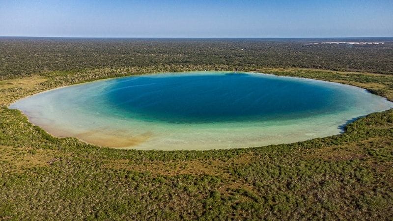 Aerial view of a large, round, blue lake surrounded by dense green forest under a clear blue sky. No notable landmarks or people visible.