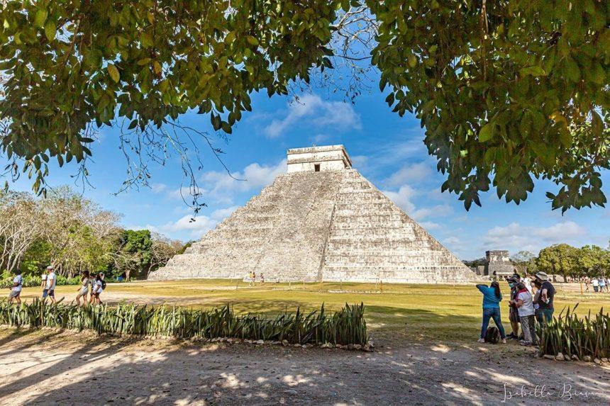 A group of people explores the ancient Mayan pyramid, El Castillo, at Chichen Itza, under a clear blue sky framed by leafy trees.