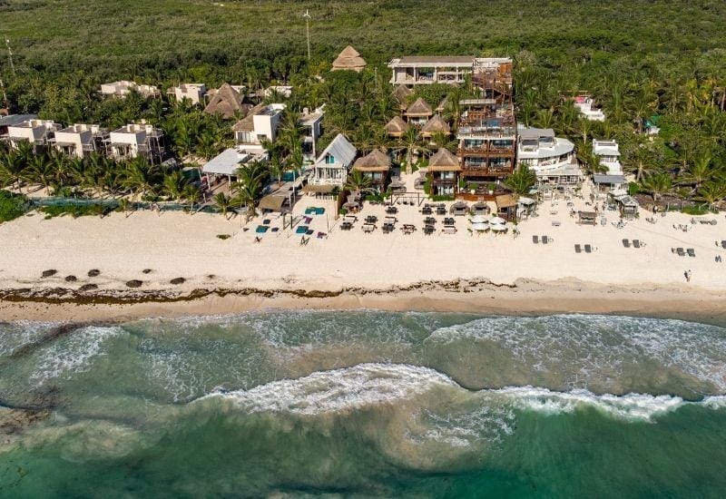 Beachfront with lounge chairs and umbrellas, bordered by dense greenery and coastal structures. The scene is tranquil with gentle waves and clear skies.