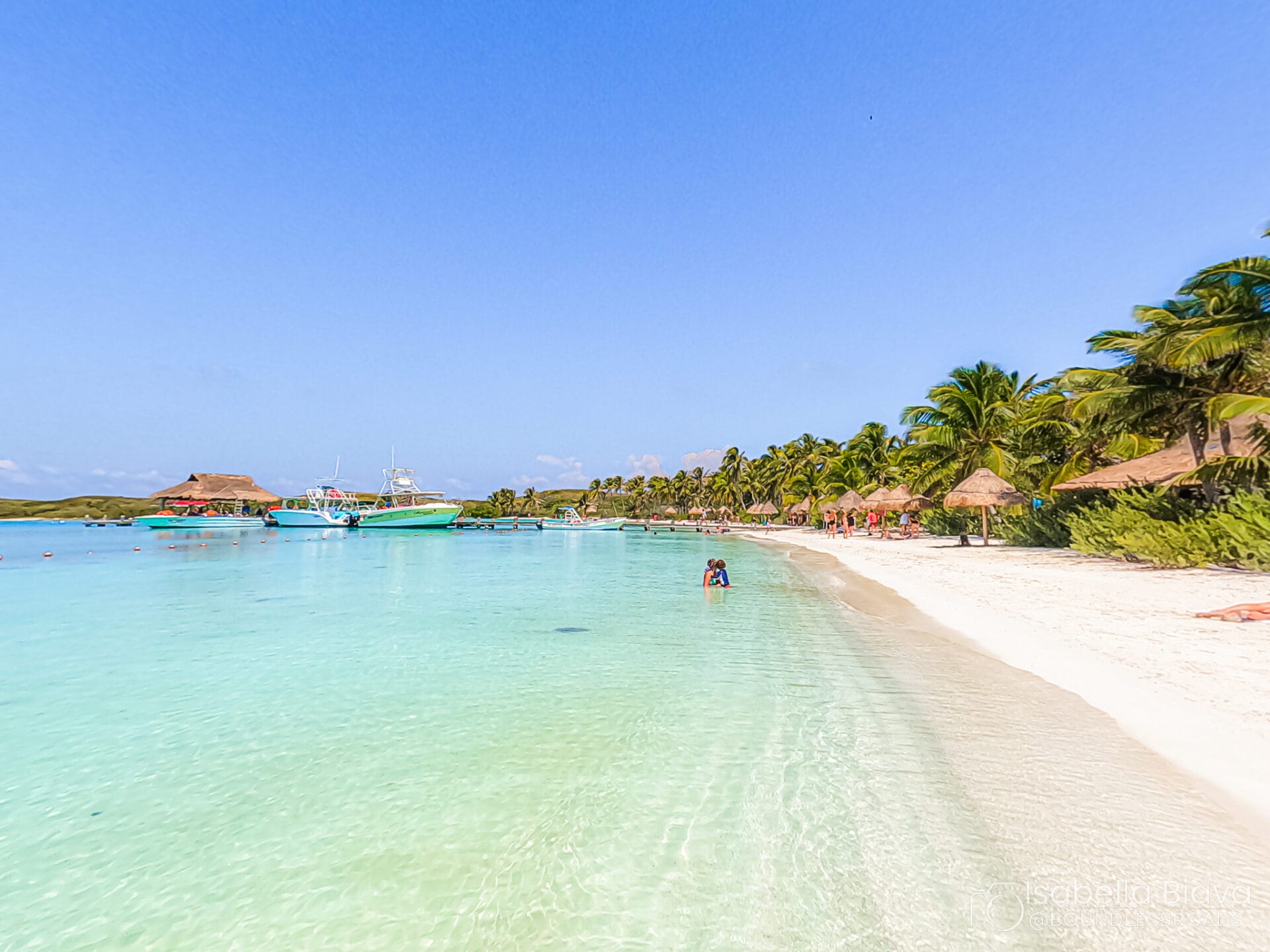 Beach scene featuring clear turquoise water, a boat, sandy shore, and palm trees. A person is in the water under a bright blue sky.