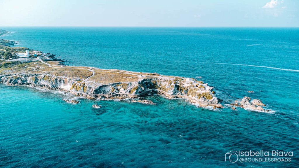 Aerial view of a rocky coastline with turquoise waters, featuring sparse vegetation. A small structure is visible on the left, surrounded by vast ocean.