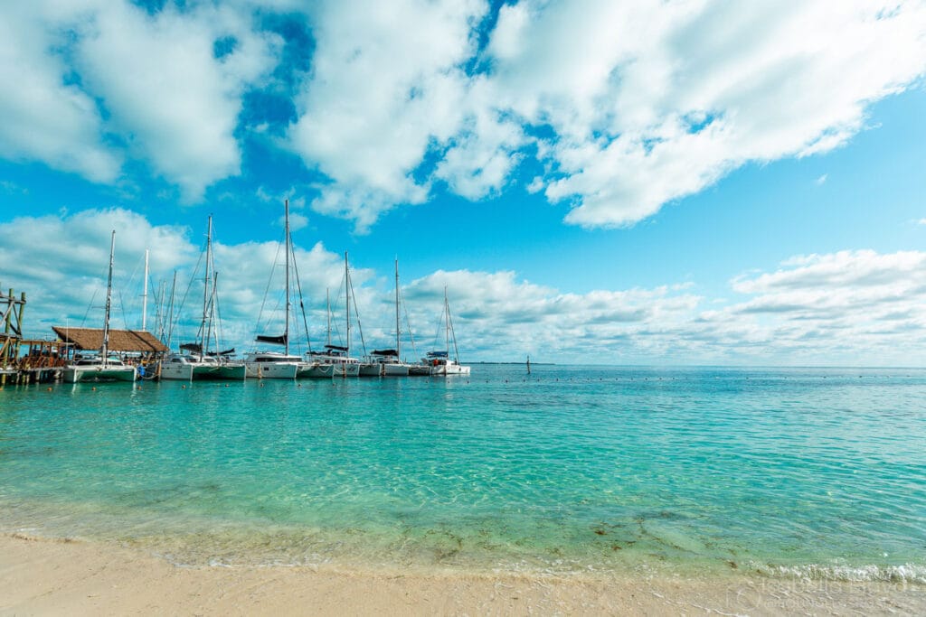 Several sailboats docked at a pier on a clear day with turquoise water and a sandy beach. The sky is filled with clouds.