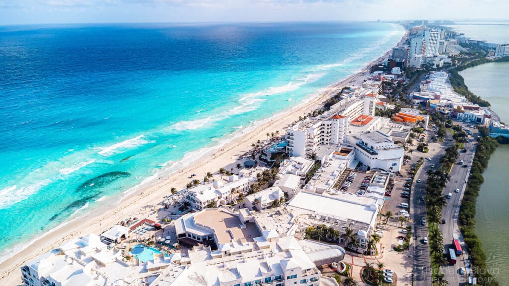 Aerial view of Cancun's beachfront with turquoise waters, hotels, and bustling streets. Sandy shores stretch alongside a vibrant cityscape.
