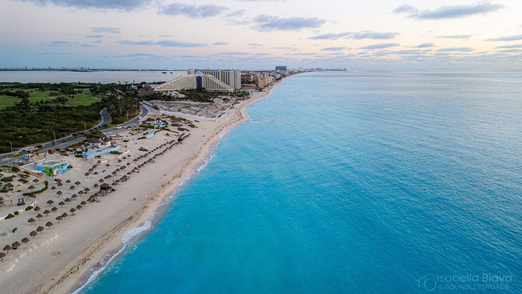 Aerial view of Cancún beach, turquoise waters, sandy shores, hotels, and lush greenery. Calm waves lap the shore, dotted with umbrellas and huts.