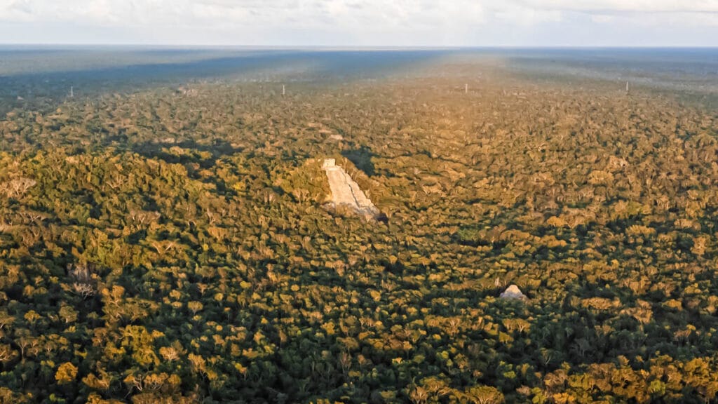 Dense forest landscape with two visible ancient structures, including the Temple of the Great Jaguar at Tikal, illuminated by sunlight under a cloudy sky.