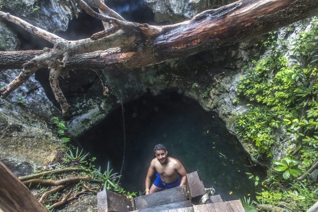 A person is climbing out of a natural cenote surrounded by rocky walls and lush greenery, with a large fallen tree above.