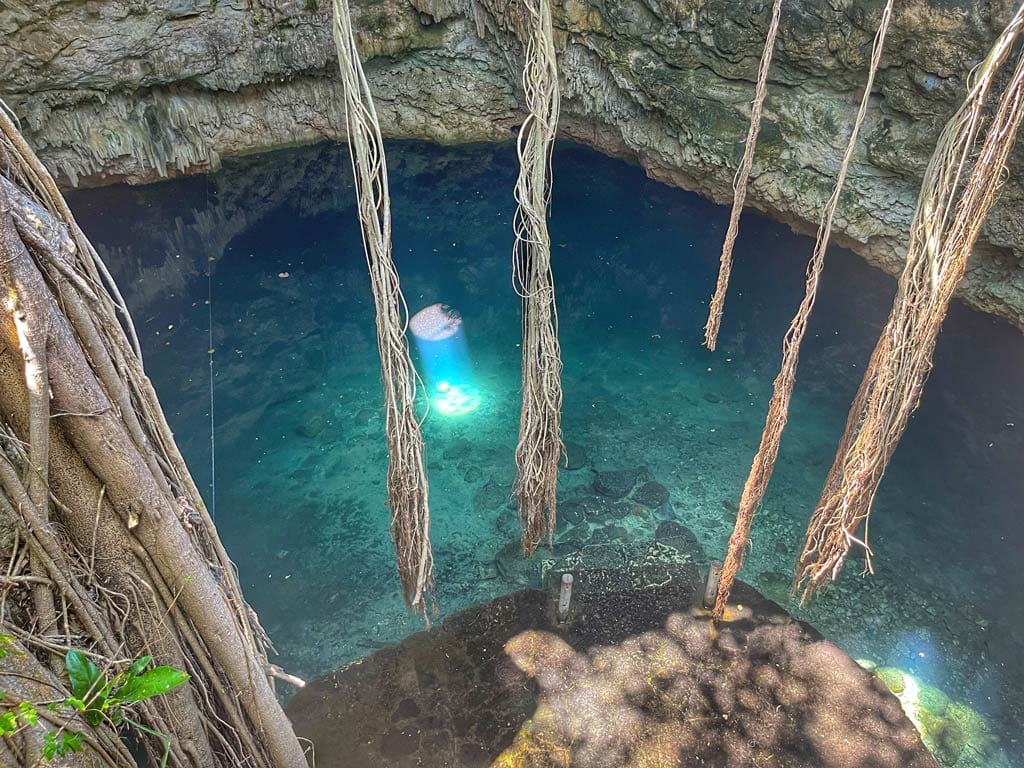 A serene cenote with clear turquoise water, surrounded by rock formations and hanging tree roots, illuminated by sunlight through the opening above.