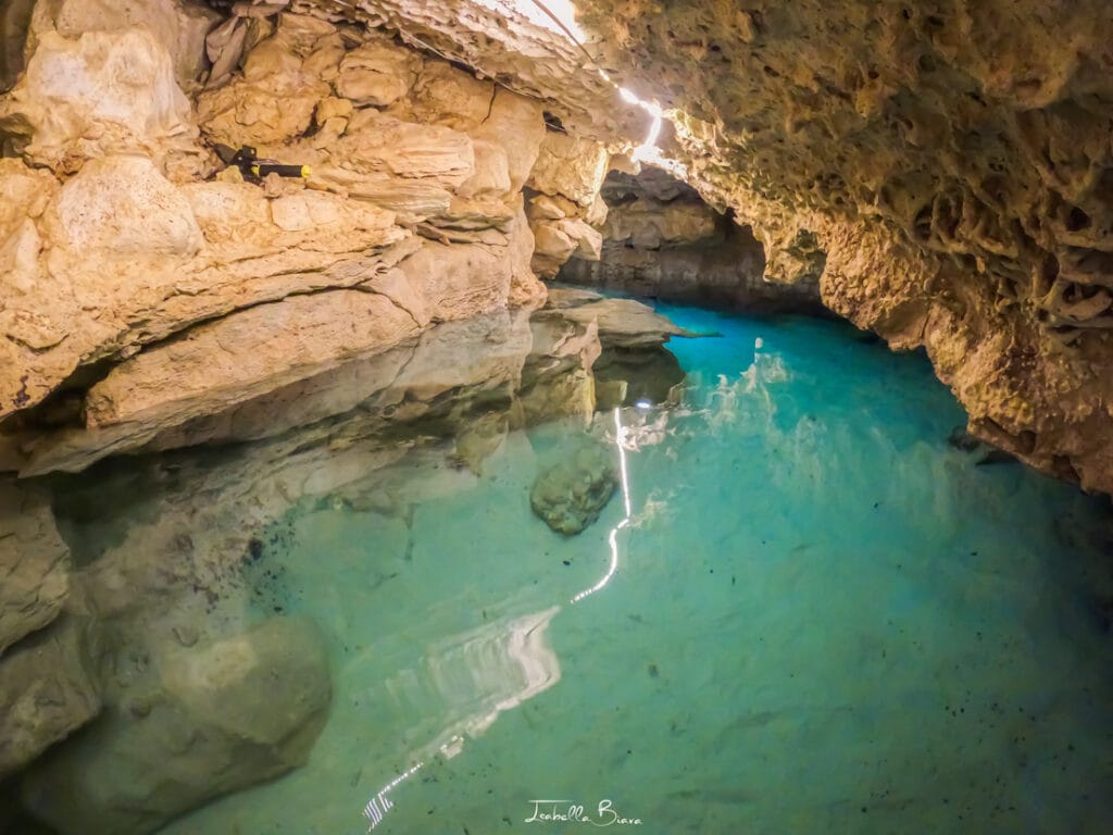 A clear turquoise pool nestled within a rocky cave, with sunlight streaming through the entrance, illuminating the water and walls.