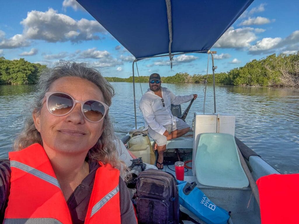 Two people on a boat in a lush, calm waterway. The person in front wears sunglasses and an orange life jacket, enjoying the scenery.