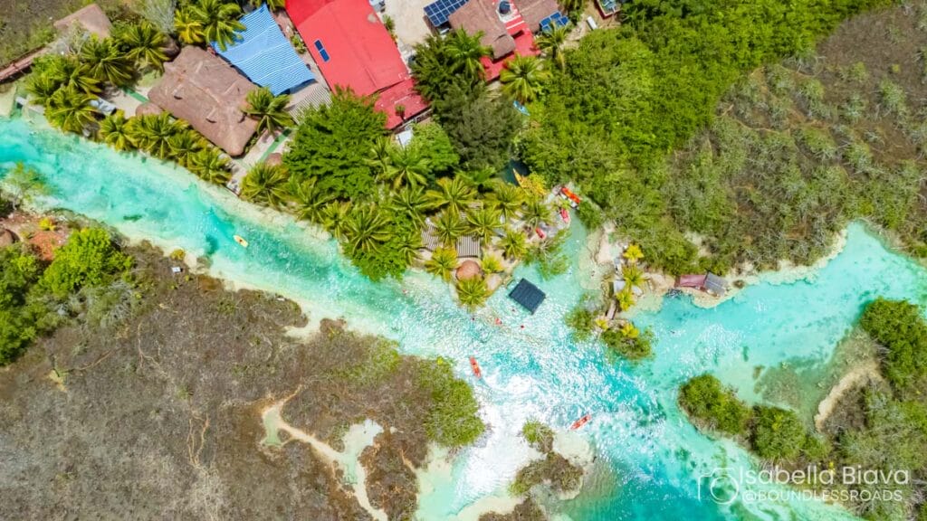 Aerial view of a turquoise river surrounded by lush greenery and buildings with red roofs; people swim and relax along the water.