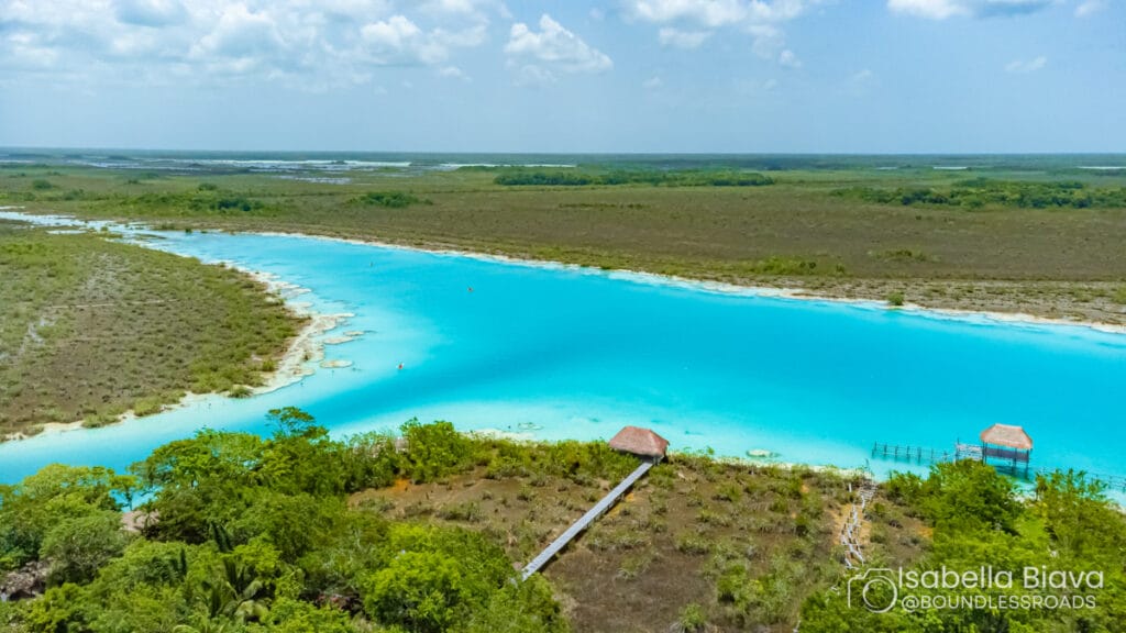 Aerial view of Bacalar Lagoon's vibrant turquoise water surrounded by lush greenery, with a wooden pier extending towards the water under a clear sky.
