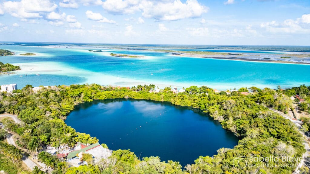 Aerial view of Cenote Azul surrounded by lush greenery, with turquoise waters of Bacalar Lagoon under a partly cloudy sky, Quintana Roo, Mexico.