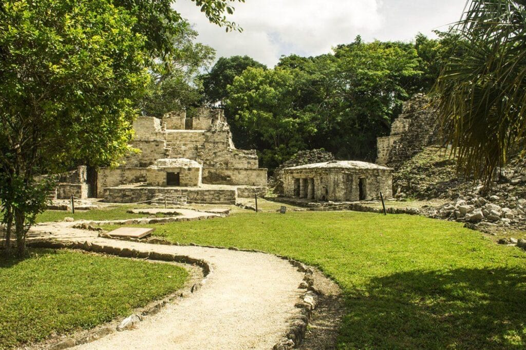 Ancient Mayan ruins at Kohunlich, Mexico, surrounded by lush greenery and stone pathways under a cloudy sky, highlighting historic architecture amidst nature.