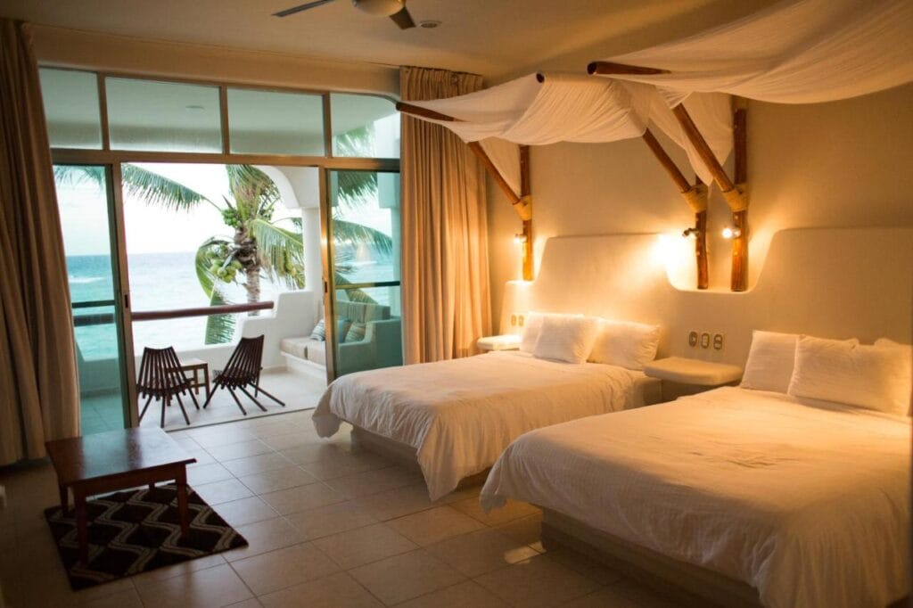 A serene hotel room with two beds, overlooking a balcony with ocean view and palm tree, featuring minimalistic decor and warm lighting.