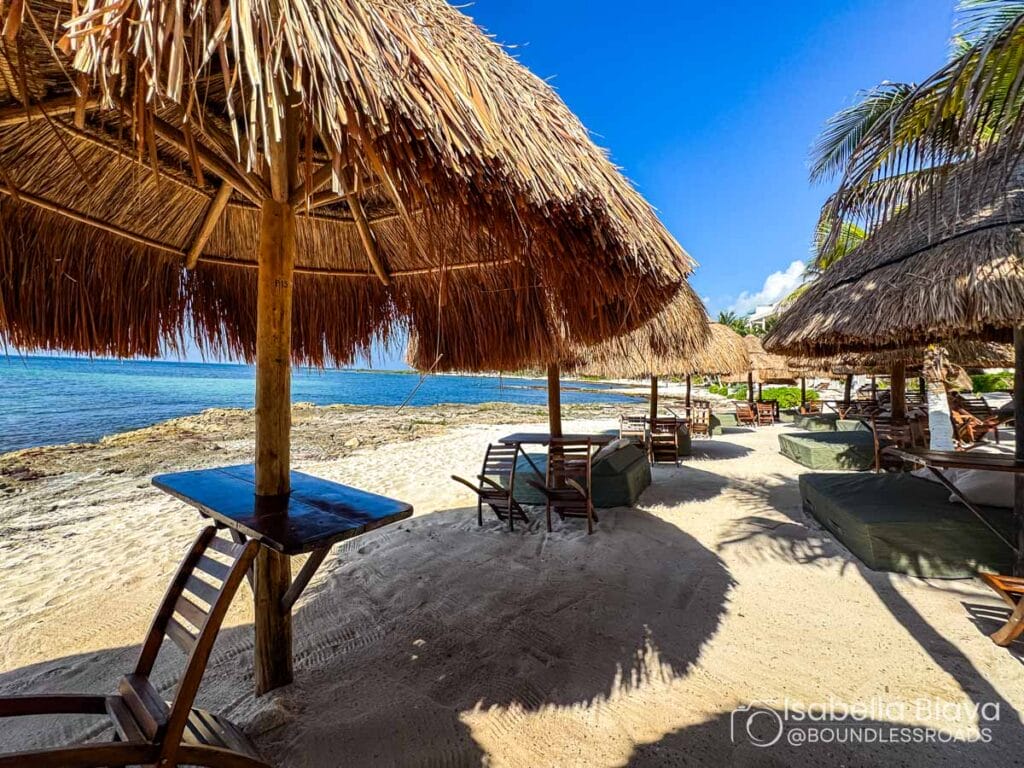 A sandy beach with thatched umbrellas, lounge chairs, and a crystal-clear ocean under a bright blue sky. No people visible.