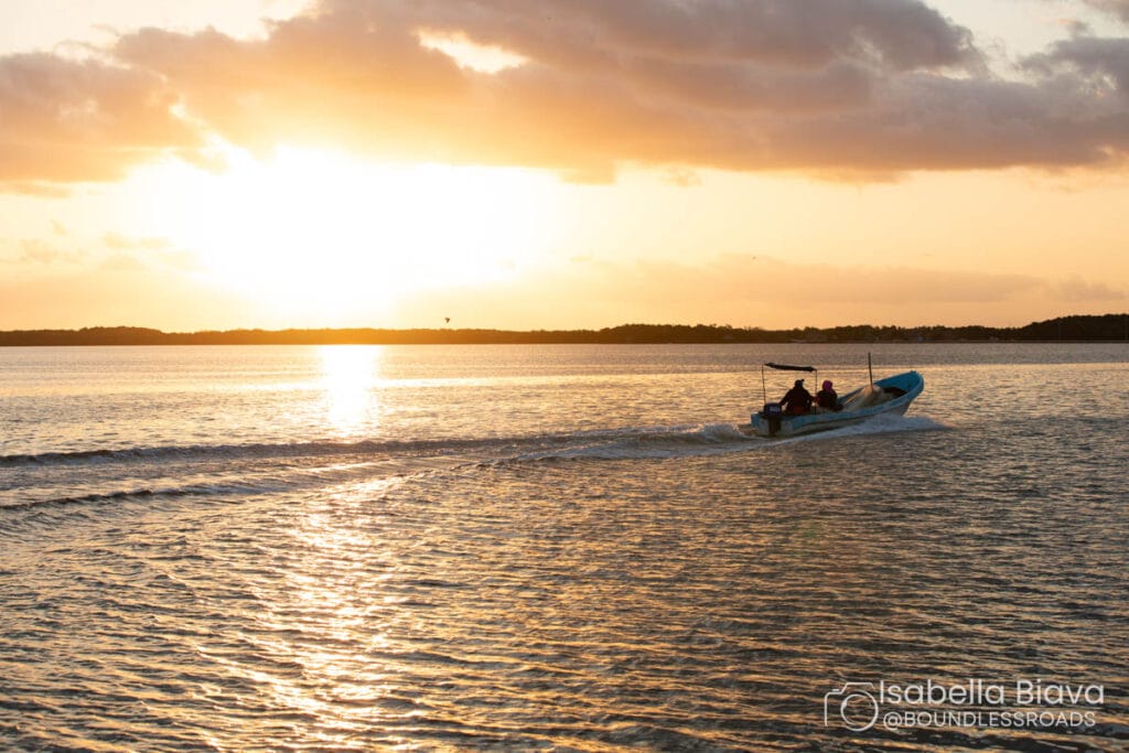 A small boat with two people moves across calm water at sunset, creating a gentle wake under a colorful sky with scattered clouds.