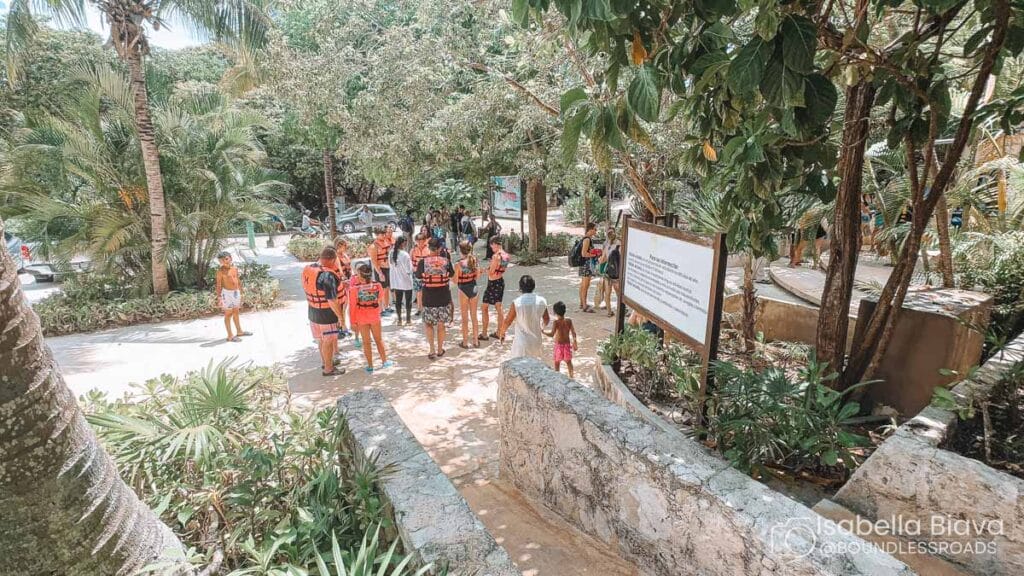 A group of people in life jackets gather near a tropical pathway surrounded by lush greenery and stone walls, with informational signage nearby.