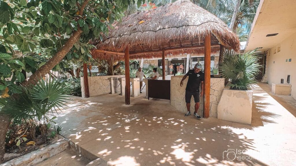 A person stands near a thatched pavilion surrounded by trees. The area appears tropical, with shaded pathways and a resort-like atmosphere.