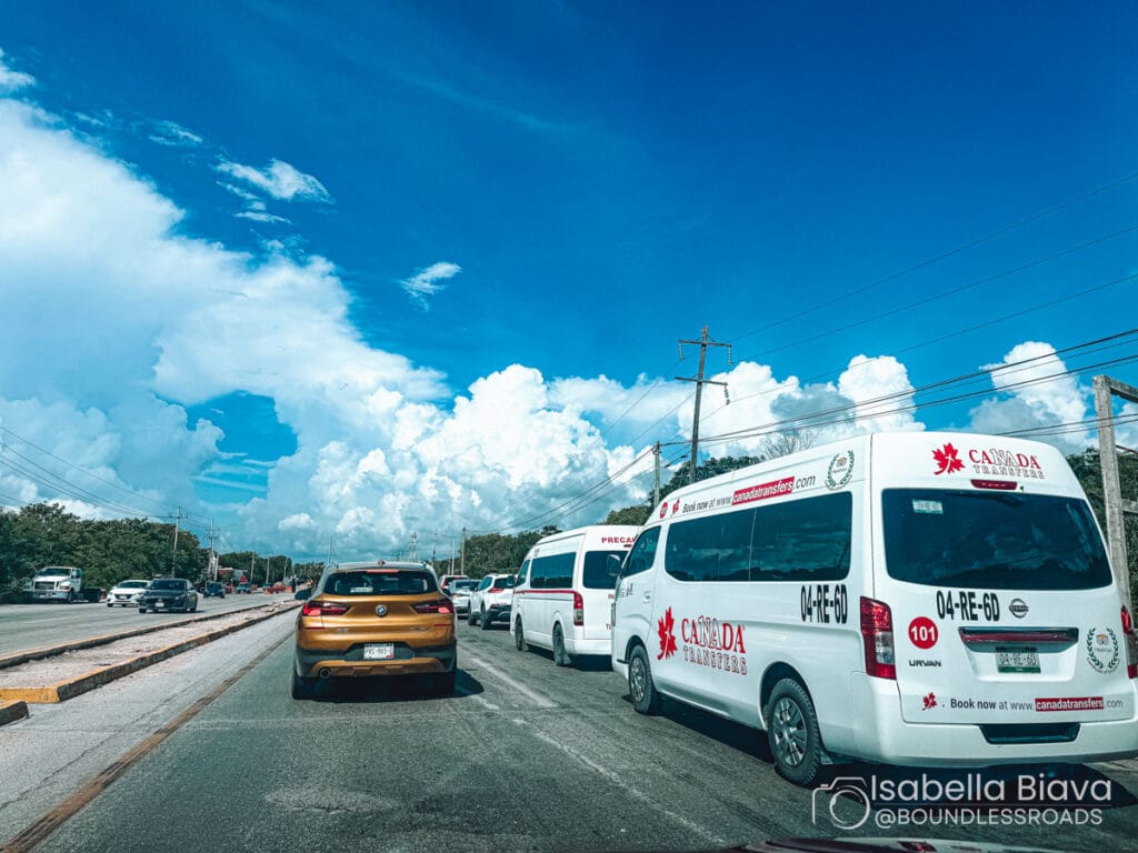 A road with vans and cars under a bright blue sky, surrounded by lush greenery and large fluffy clouds. No recognizable landmarks.