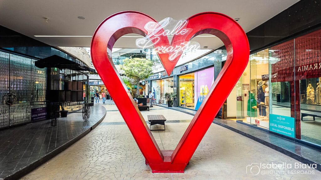 A large red heart sculpture with "Bialoleka" in neon stands in a shopping mall passage, surrounded by various retail stores.