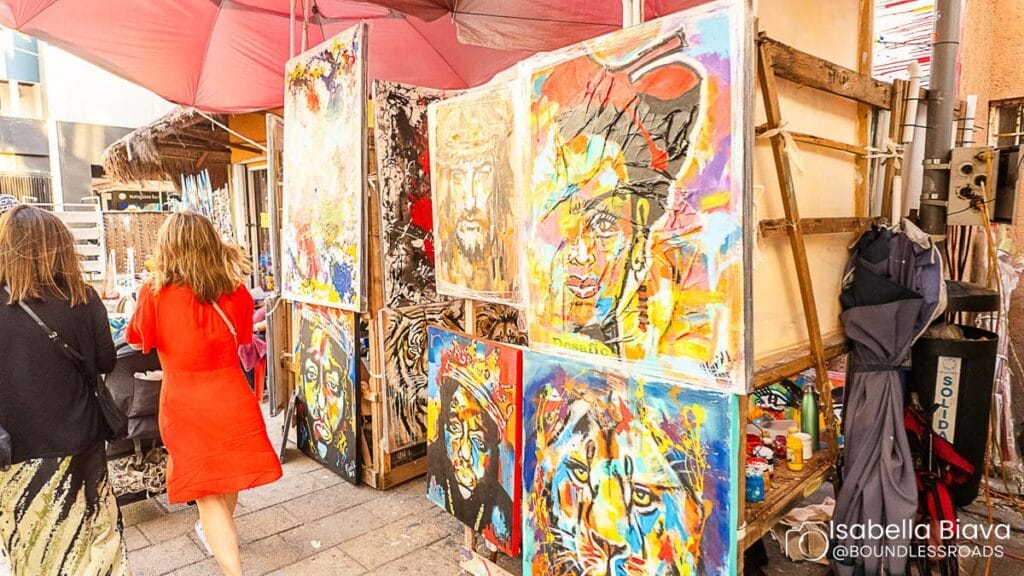 Two people walk past a vibrant street art display featuring colorful paintings under umbrellas. Urban setting with no identifiable landmarks or buildings.
