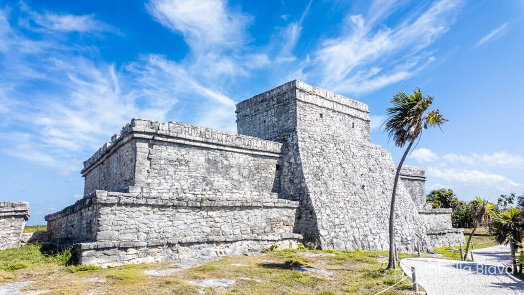 Ancient stone structure at Tulum ruins under a clear blue sky with palm trees nearby. Historical architecture blends with natural beauty.