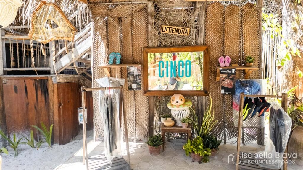 Rustic outdoor beach shop with tropical decor, featuring a sign for "Cinco" and various goods displayed on wooden surfaces, surrounded by lush greenery.