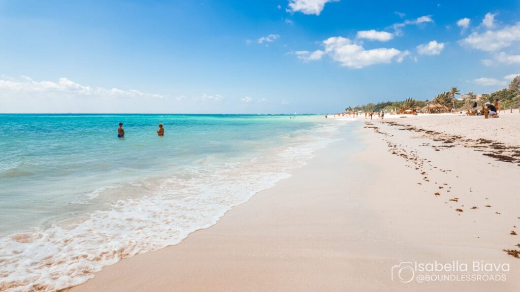Sunny beach scene with turquoise water and white sand. Two people in the sea, distant beachgoers, palm trees, and clear blue sky.