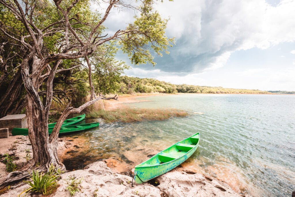 A scenic lakeside view with green canoes on the rocky shore, surrounded by trees and under a partly cloudy sky.