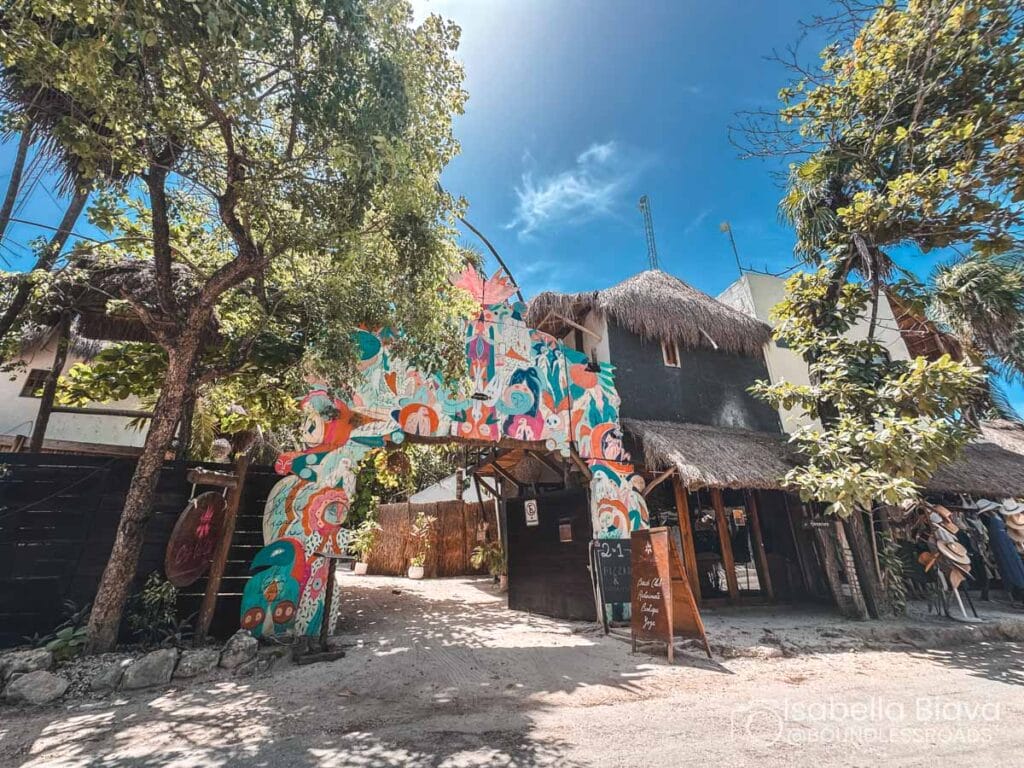 Colorful, tropical entrance to a rustic establishment with thatched roofs and lush trees, under a vibrant blue sky. No recognizable landmarks or historical buildings.